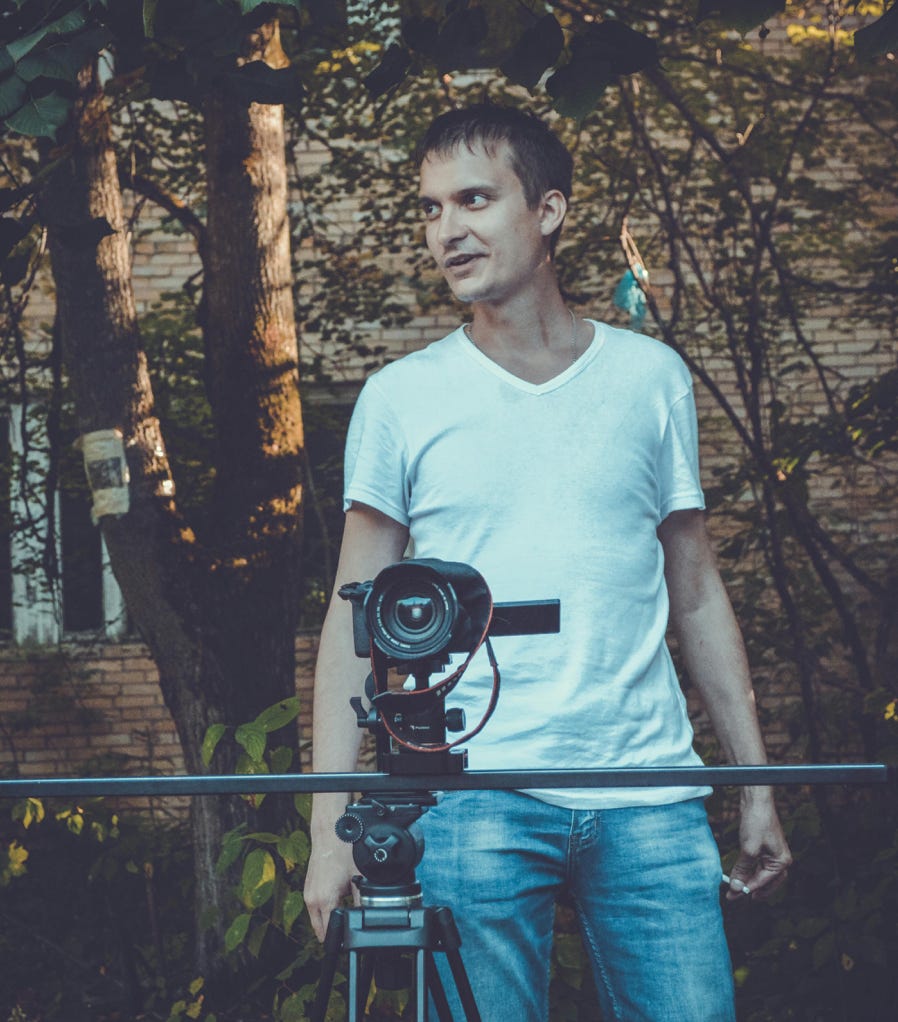 Camera man in white shirt and jeans looking away while taking video on professional equipment on tripod
