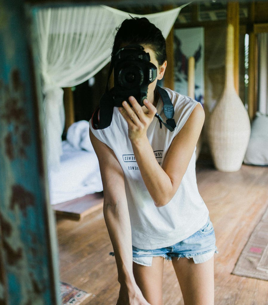 Mirror reflection of unrecognizable young slender female in white t shirt and denim shirts taking selfie in bright room with old weathered shabby walls